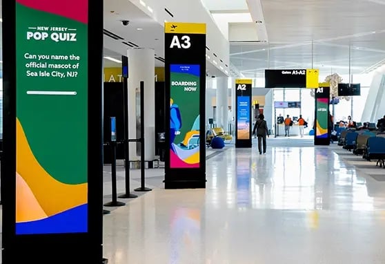 Airport travelers walking past a row of triangular digital pylons with dual LED faces displaying flight information, boarding cues and New Jersey trivia.