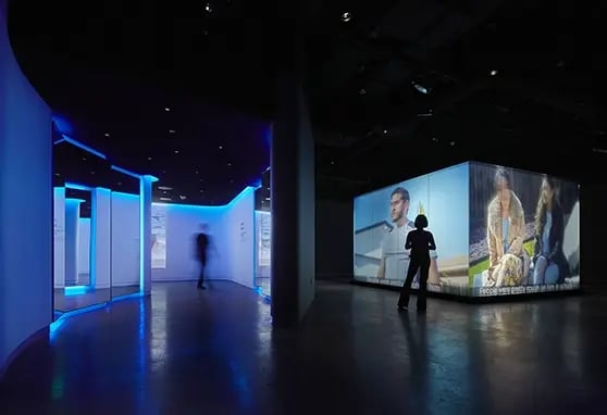 A dark exhibition space features illuminated blue walls and large screens displaying videos, with a silhouette of a visitor in motion.