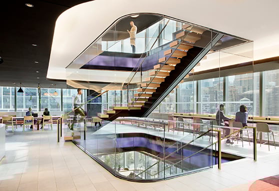 A dynamic and open office environment. The centerpiece is a sculptural staircase encased in glass and LED walls, visually connecting multiple floors and encouraging fluid movement and interaction. Subtle lighting accents and warm wood tones contribute to an inviting atmosphere, while panoramic city views highlight the urban context.