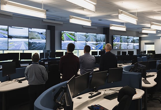 A group of people monitors multiple live traffic camera feeds in a modern traffic control center equipped with numerous workstations and large display screens.