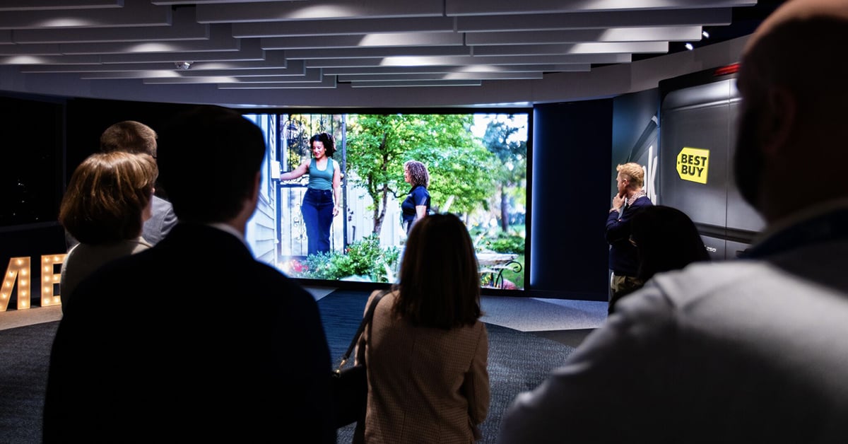A group of people observe a dynamic video presentation in a modern showroom environment featuring immersive audiovisual technology and branded content.