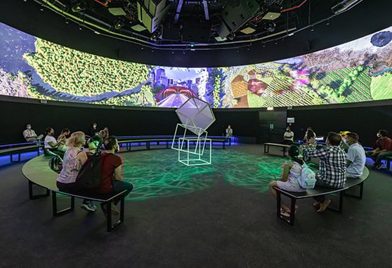 A group of people sits on benches in a dark circular room with a digital screen displaying nature scenes above them and a geometric sculpture in the center.