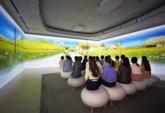 A group of people sits on round white stools in a modern immersive theater, watching a vivid animated projection that spans three walls, depicting a bright rural scene with grassy fields, huts, and a winding road.