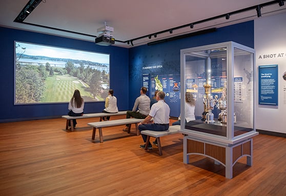 A group of visitors sits on benches in a modern museum exhibit space, watching a projected video of a scenic golf course. The room features informative wall displays about golf tournaments and a glass case showcasing prestigious golf trophies. The exhibit appears to focus on the history and planning of major golf championships, creating an engaging and educational experience.