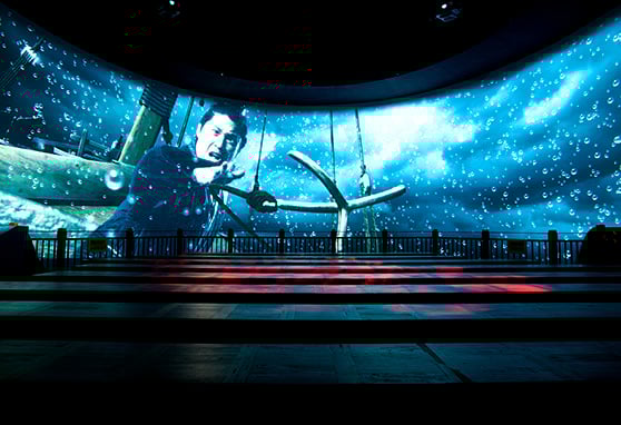 A large immersive curved screen displays a dramatic underwater scene with a man reaching out while gripping a large anchor. The background features a stormy ocean. In front of the screen are tiered wooden platform seating.