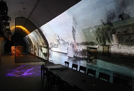 A long, arched tunnel at the Dover War Tunnels with historical images of steamships and riverscapes projected across a curved brick wall, creating an immersive environment. A dark table and chairs line the center.