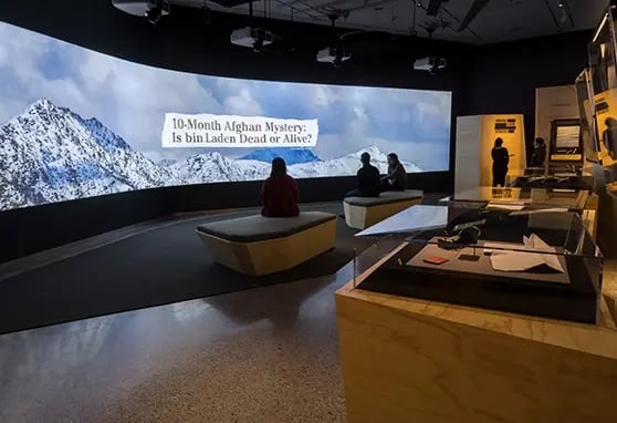 A modern exhibit space featuring a large screen displaying a question about Osama bin Laden, with visitors engaged in discussion.