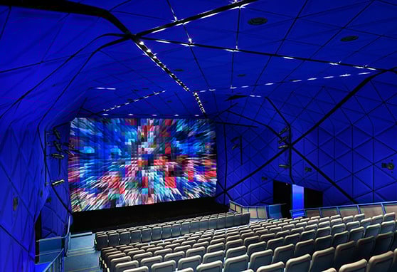 A modern theater with blue geometric walls, empty gray seats and a large screen displaying a blurred, colorful abstract image.