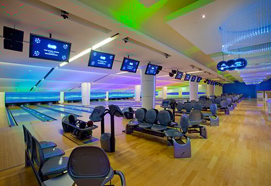A modern, high-end bowling alley with a colorful and vibrant atmosphere. The setup is sleek, with smooth wood flooring, ergonomic seating, and automated scoring monitors above each lane. The lighting is a standout feature—LED strips and ceiling lights glow in shades of blue, green, and purple, giving the space a futuristic, club-like vibe.