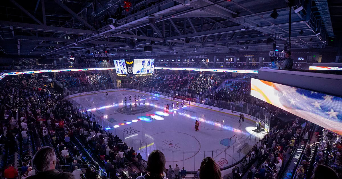 A packed hockey arena buzzes with energy as fans stand for the national anthem before the start of a game. The rink is illuminated with patriotic lighting effects, casting red, white, and blue across the ice. A massive scoreboard hangs above center ice, displaying player visuals and game details, while a digital ribbon board wraps around the arena showcasing an American flag. The atmosphere captures the excitement and unity of a major sporting event in a modern, high-tech venue.