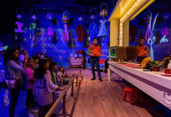 A performer holding oversized carrots interacts with a group of children at an immersive, theatrical exhibit. The space is colorfully lit with purple and blue hues, and features whimsical costumes hanging from the ceiling.