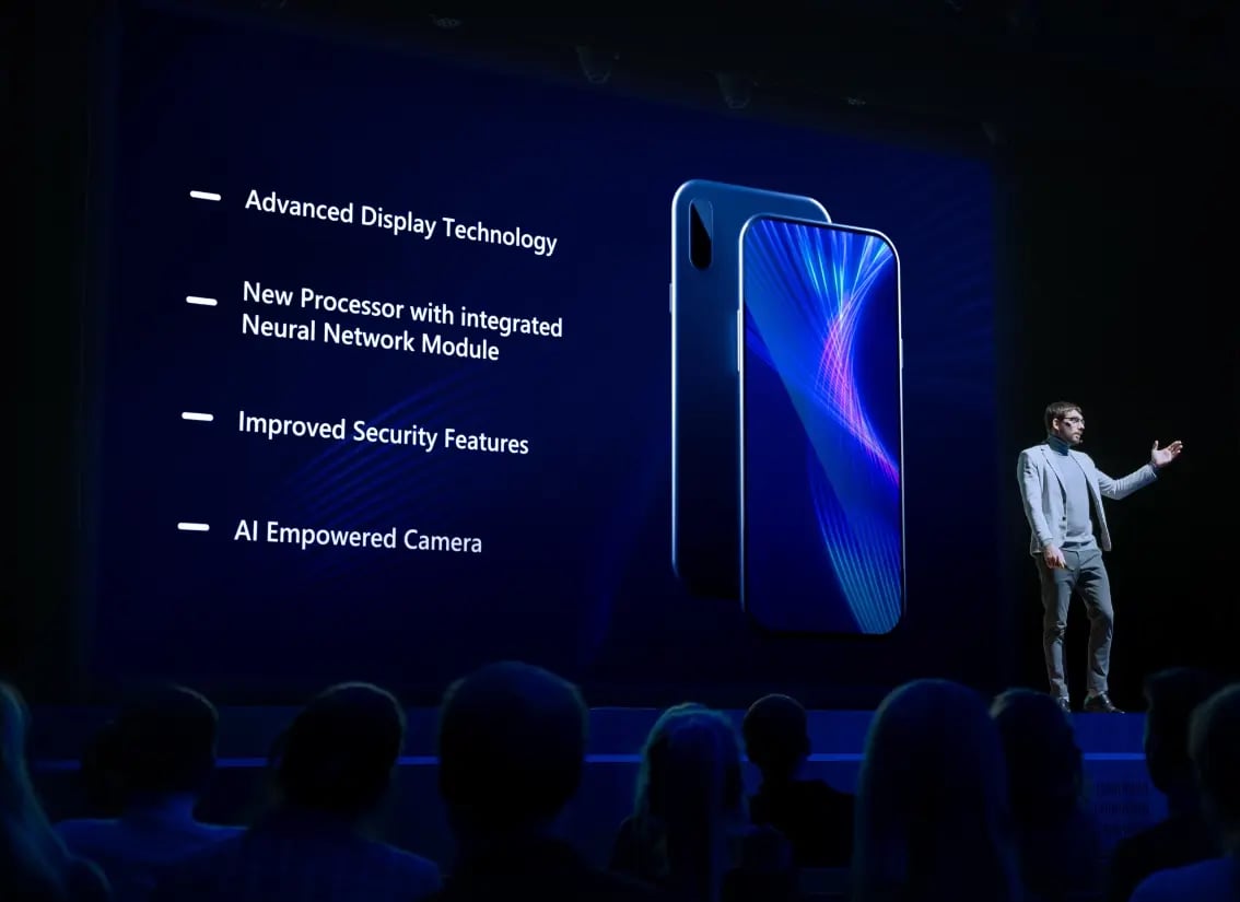 A presenter at a product launch talking to the audience while information about the product is shown behind on a large screen.