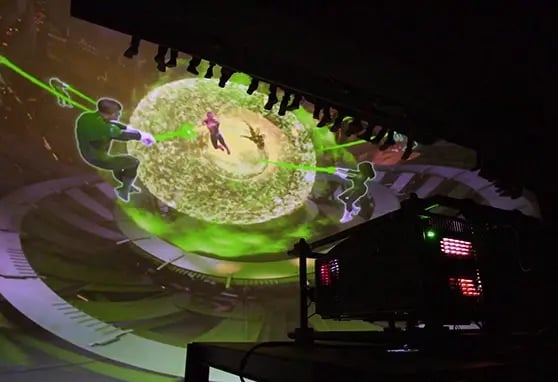 A projection of two figures with green lasers facing a central figure, surrounded by a glowing, swirling sphere in a dark setting. Below is a projector.