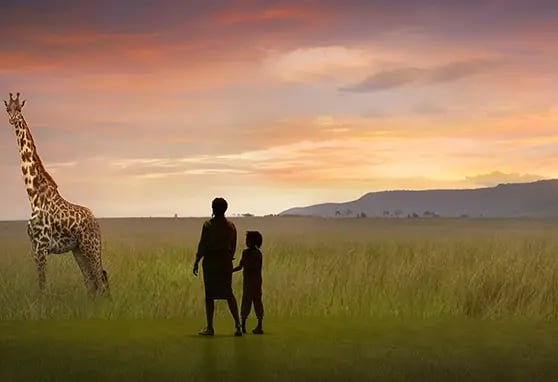 A silhouetted adult and child stand in a projection mapped grassy field, gazing at a giraffe under a colorful sunset sky.