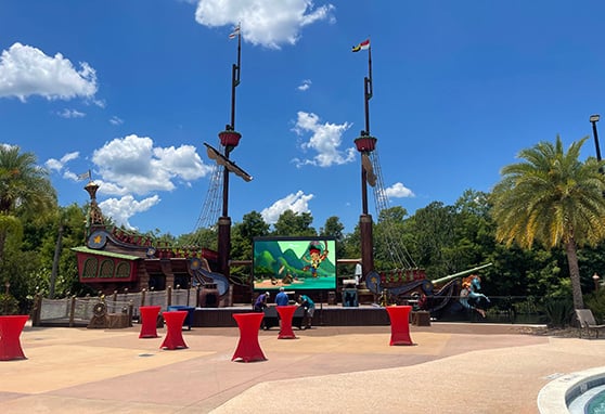 A whimsical, pirate-themed play area or entertainment stage that looks like it's part of a family resort or amusement park. The main attraction is a large, colorful pirate ship structure—complete with masts, sails, rigging, and cartoonish details—flanked by palm trees and surrounded by lush greenery under a bright, sunny sky.