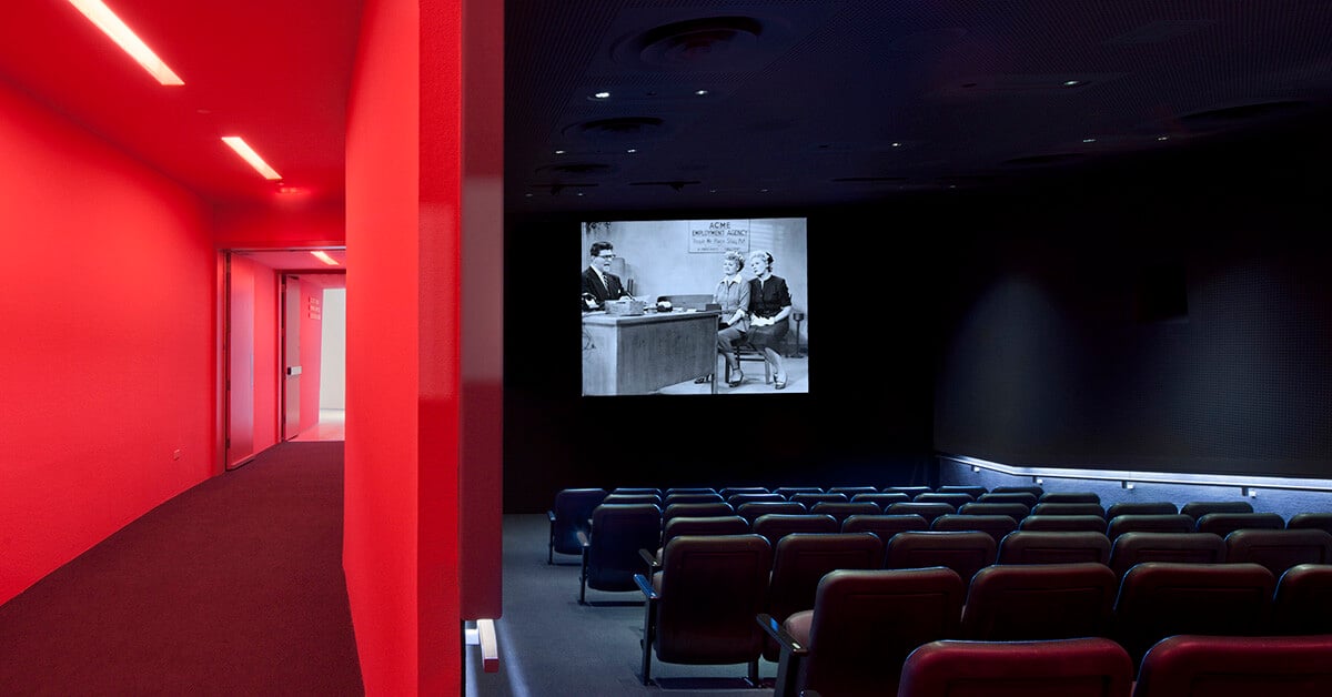 A modern cinema room with rows of empty, dark red theater seats facing a screen playing a black-and-white film. The right side of the image shows the dark, intimate interior of the theater, while the left side reveals a bright red hallway.