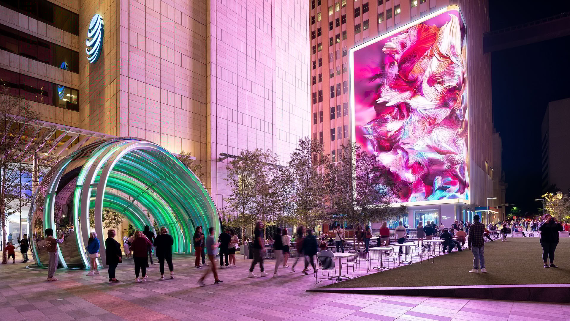 The vibrant AT&T Discovery District. The scene is anchored by a striking, oversized LED display with swirling pink and white visuals on the side of a tall building. A glowing AT&T Globe installation attracts visitors nearby, while people stroll, gather and relax throughout the illuminated space.