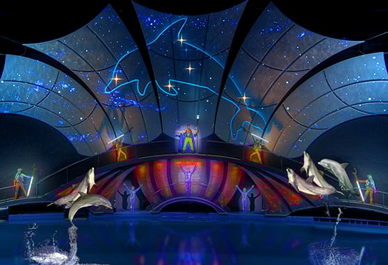 A vibrant aquatic stage show featuring dolphins leaping from a pool in front of a futuristic, starry backdrop. Performers in colorful costumes stand on elevated platforms infront of a curved, sail-like video displaying a celestial scene.