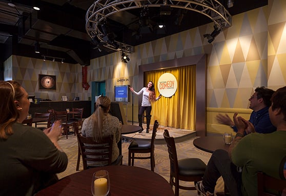 A woman performs on a small stage at the National Comedy Center while an audience seated at café-style tables applauds. The room features geometric-patterned walls, a circular lighting truss above the stage, and a cozy, intimate comedy club atmosphere.