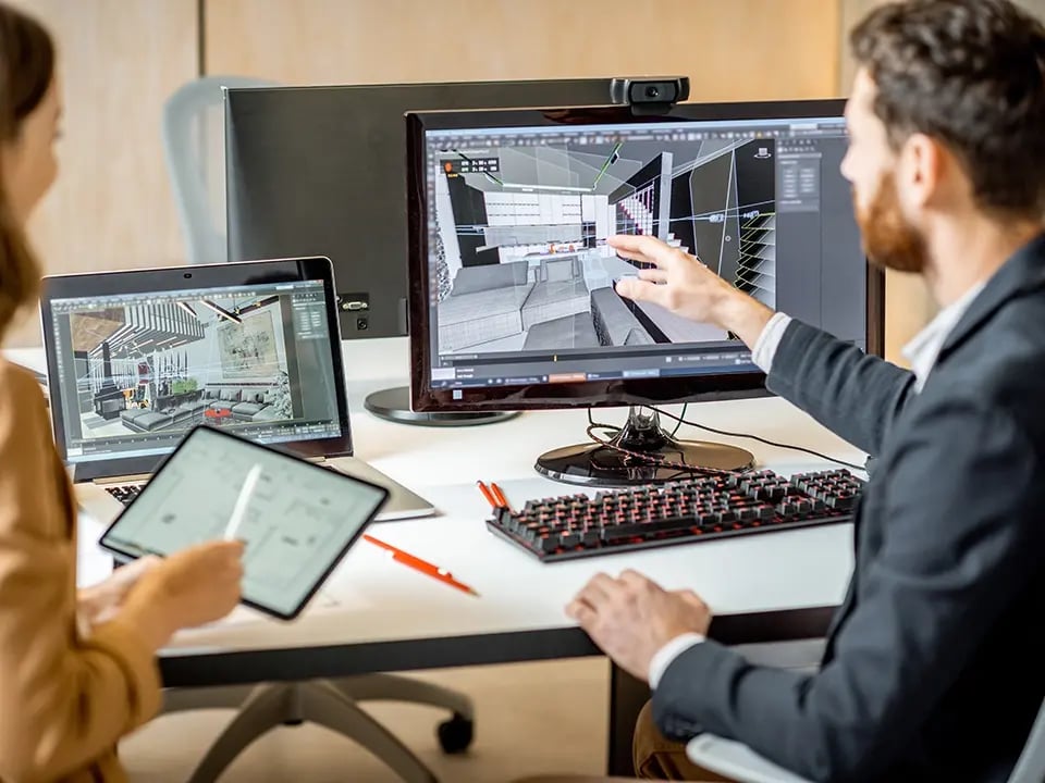 A man and a woman collaborate at a desk, working on 3D interior design models displayed on a computer monitor and a laptop screen. The man is pointing at the desktop screen showing a wireframe layout of a modern living room, while the woman holds a tablet and stylus, reviewing design elements. The workspace appears modern and professional, suggesting a design or architectural studio.