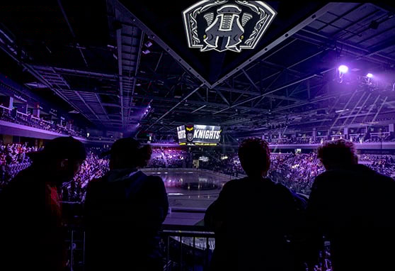 Crowd gathers at a packed indoor arena with dramatic lighting and a large central screen displaying 'Knights' at Dollar Loan Center; silhouetted fans watch from the balcony as the ice rink below reflects purple hues and team branding.