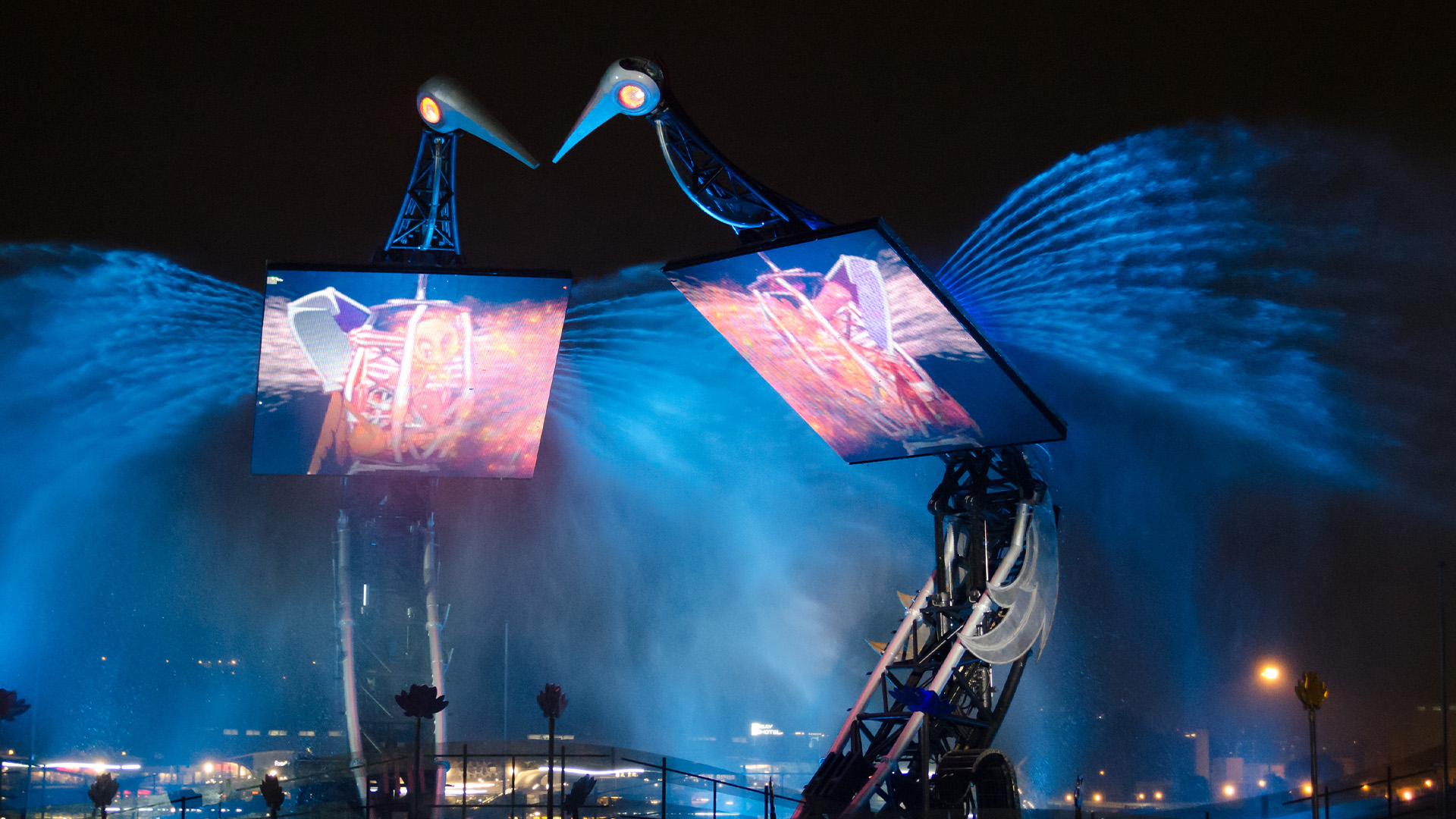 A visually stunning nighttime spectacle involving two giant mechanical bird-like structures, which are part of a multimedia installation or performance. The two large screens mounted on the structures display vibrant animations or videos, while synchronized water sprays arc dramatically from each "wing," illuminated in deep blues that enhance the ethereal, futuristic atmosphere.