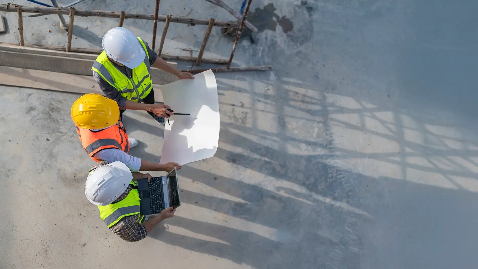 Three construction workers, wearing safety vests and hard hats, are seen from above reviewing blueprints and a laptop on a concrete site. One worker holds a large sheet of paper while another gestures with a pen or pointer, and the third consults a laptop. Shadows of the workers and a nearby structure stretch across the concrete ground, indicating strong sunlight.