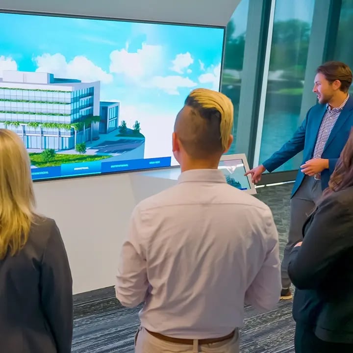 A group of professionals is gathered in a modern office space, viewing a digital architectural rendering of a building on a large screen. One man, standing beside the display, is pointing at the screen while holding a tablet, appearing to lead the presentation. The digital model showcases a contemporary multi-story building surrounded by greenery, labeled with options like 