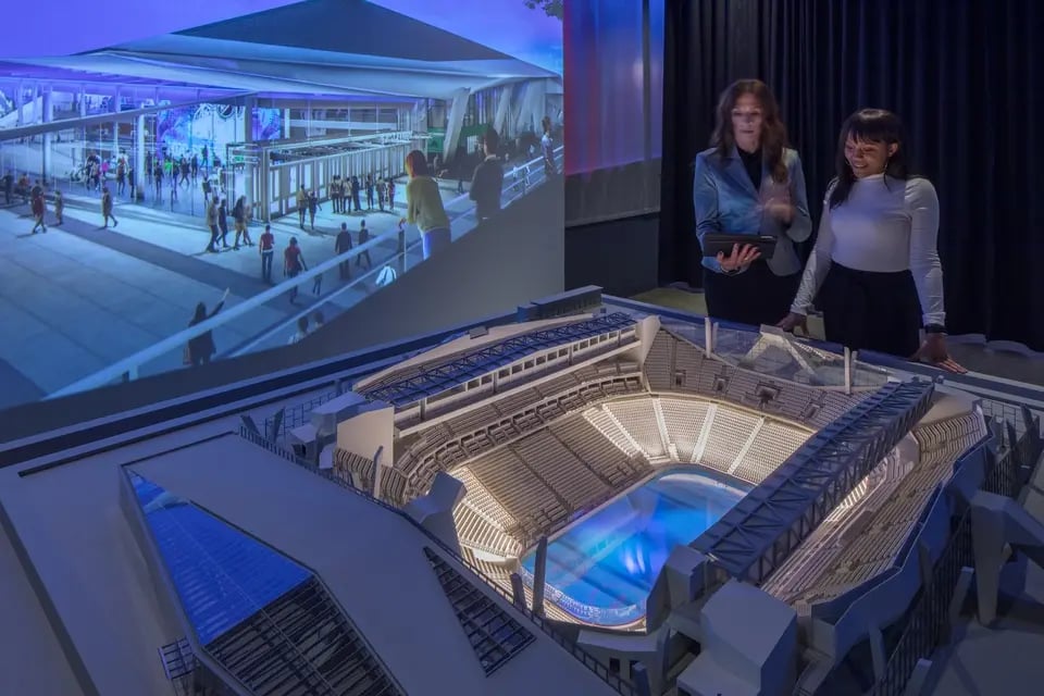 Two women examine a detailed model of a stadium paired with projection mapping solutions, discussing its technology, design and features.