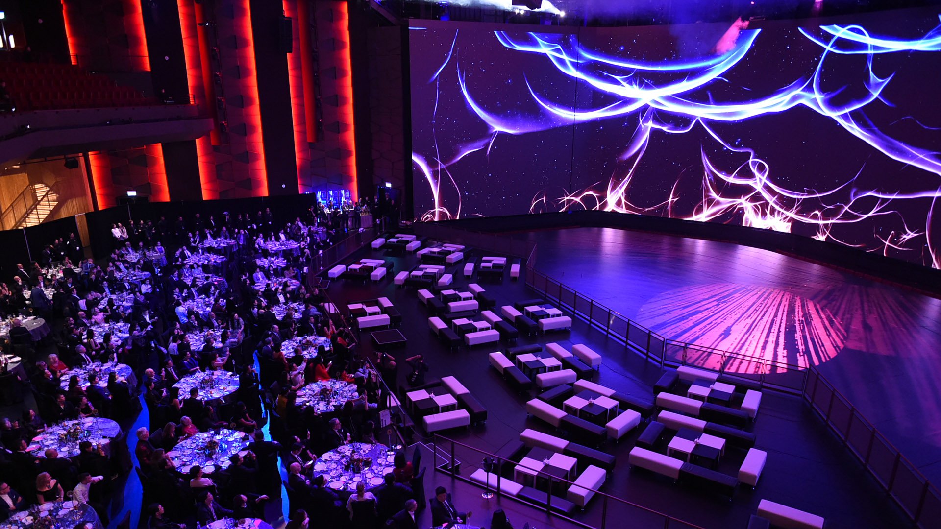 Elegant event setup inside a modern venue with round banquet tables filled with guests on the left, lounge-style seating on the right, and a massive LED screen displaying abstract  light patterns above a circular stage at Asia's first dynamic theater.