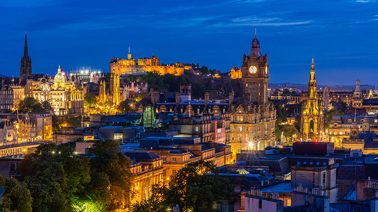 The Edinburgh skyline at dusk features the historic Edinburgh Castle perched atop Castle Rock, softly illuminated against a deepening sky. Surrounding rooftops and spires are silhouetted in the fading light, capturing the city's timeless and dramatic character.