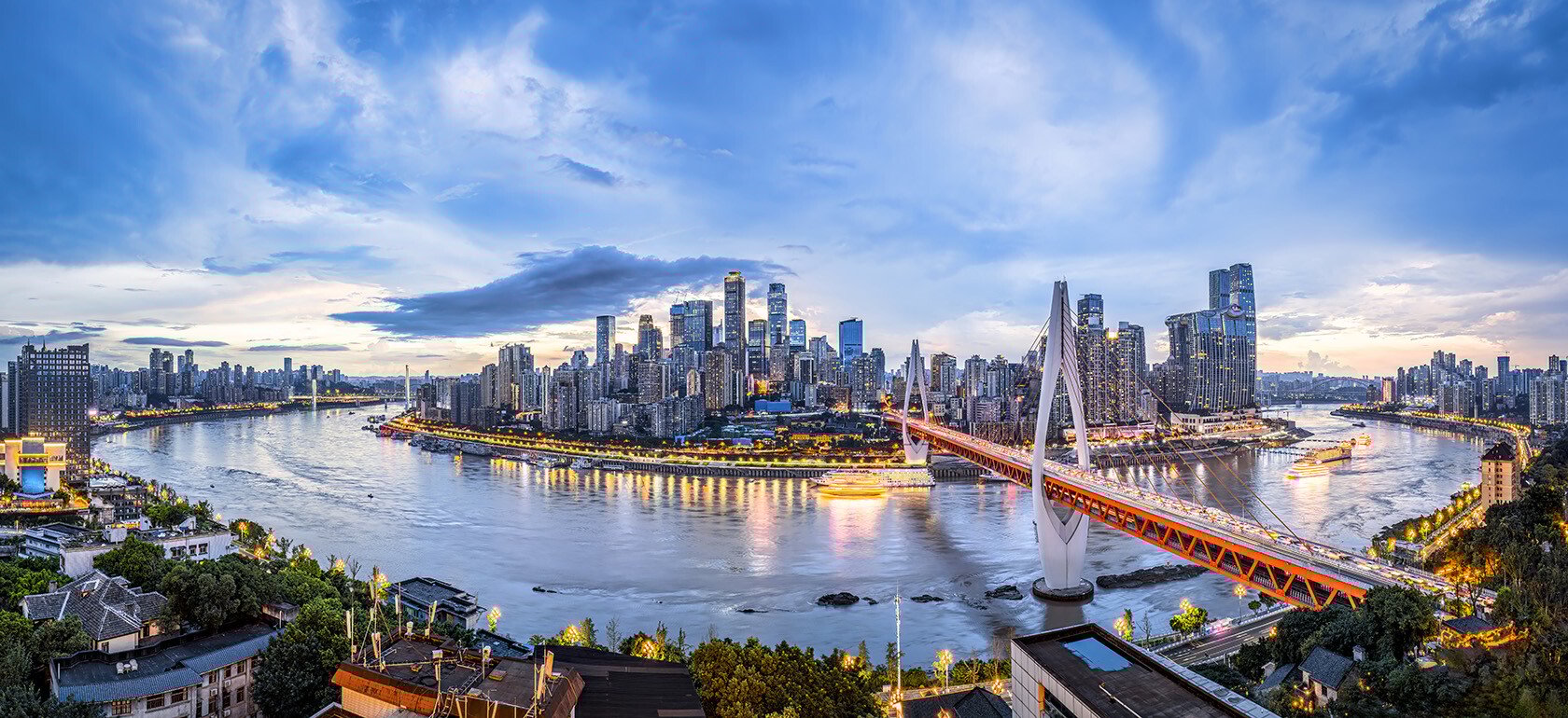 A modern city skyline at dusk glows with the soft transition of daylight to evening, as building lights begin to twinkle against a colorful, fading sky. The mix of glass towers and urban architecture reflects off nearby water, creating a serene yet vibrant cityscape.