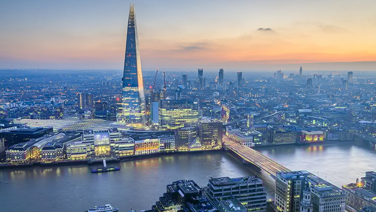 The London skyline at early dusk is softly lit with the fading daylight, highlighting The Shard as it rises prominently above the city. Warm lights begin to glow from surrounding buildings, reflecting gently off the Thames and adding a serene ambiance to the urban landscape.