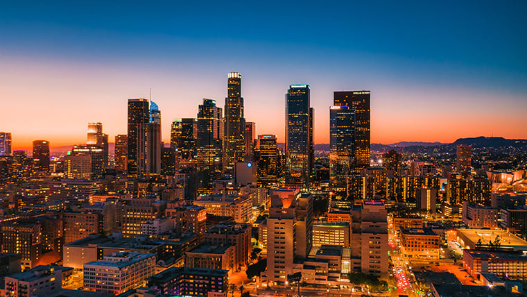 Downtown Los Angeles at dusk is bathed in a warm, golden light as the sun sets behind the skyline, casting long shadows across the city. Skyscrapers begin to glow with interior lights, while traffic and activity add movement to the urban landscape.