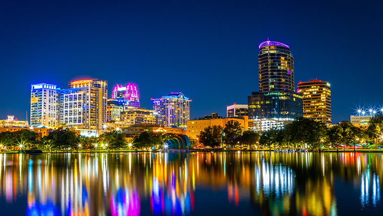 Downtown Orlando at night sparkles with illuminated high-rises and colorful lights reflecting off nearby Lake Eola. The vibrant cityscape captures the energy of nightlife and the charm of the urban skyline.