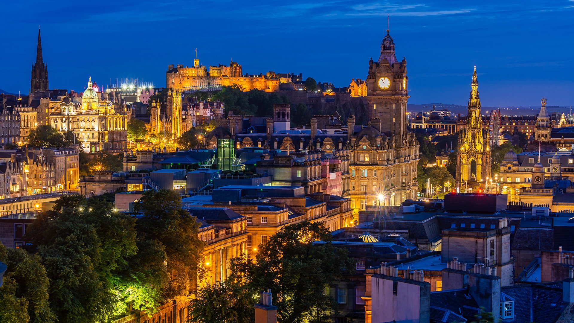 The Edinburgh skyline at dusk features the historic Edinburgh Castle perched atop Castle Rock, softly illuminated against a deepening sky. Surrounding rooftops and spires are silhouetted in the fading light, capturing the city's timeless and dramatic character.