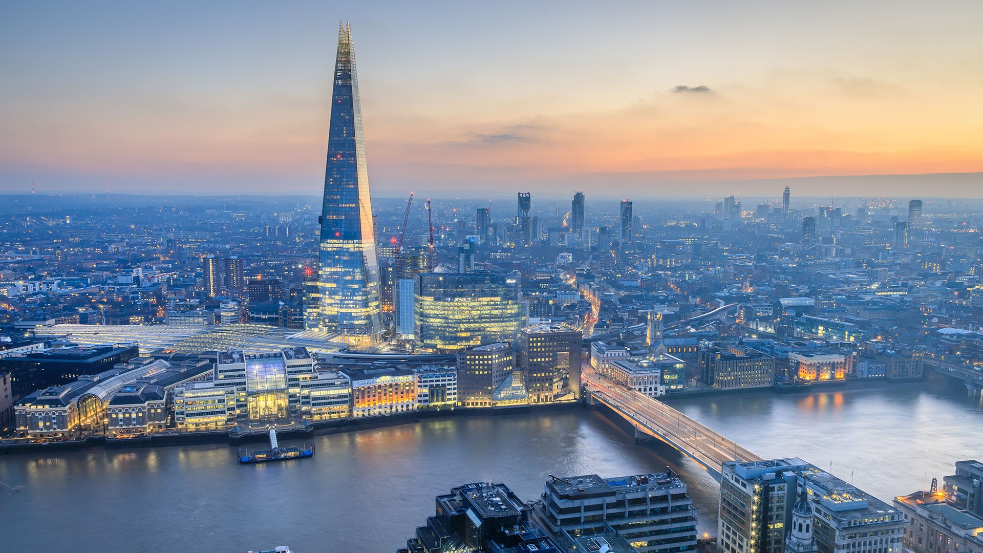 The London skyline at early dusk is softly lit with the fading daylight, highlighting The Shard as it rises prominently above the city. Warm lights begin to glow from surrounding buildings, reflecting gently off the Thames and adding a serene ambiance to the urban landscape.