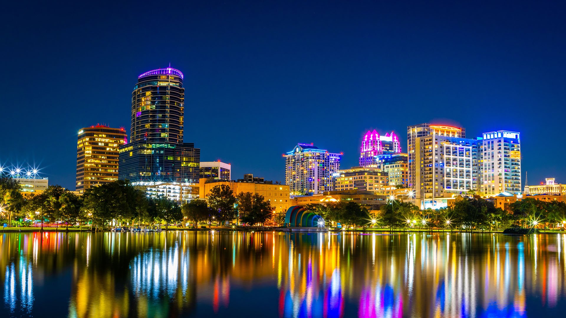 Downtown Orlando at night sparkles with illuminated high-rises and colorful lights reflecting off nearby Lake Eola. The vibrant cityscape captures the energy of nightlife and the charm of the urban skyline.