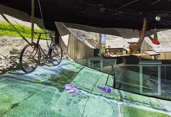 Exhibit featuring a bicycle suspended in front of angled surfaces displaying projection mapped content, with architectural models and objects in the background.