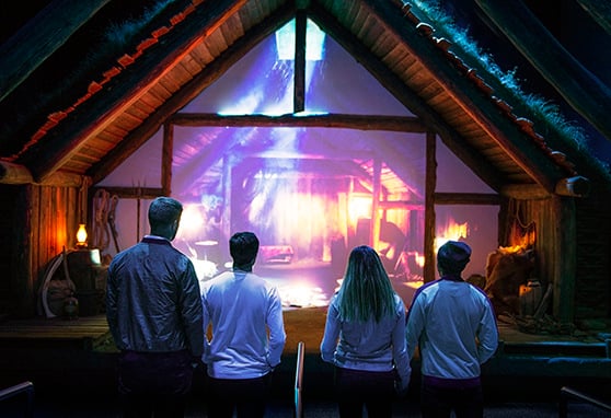 Four people stand in front of a lifesized, immersive digital display designed to look like the interior of a rustic wooden cabin.