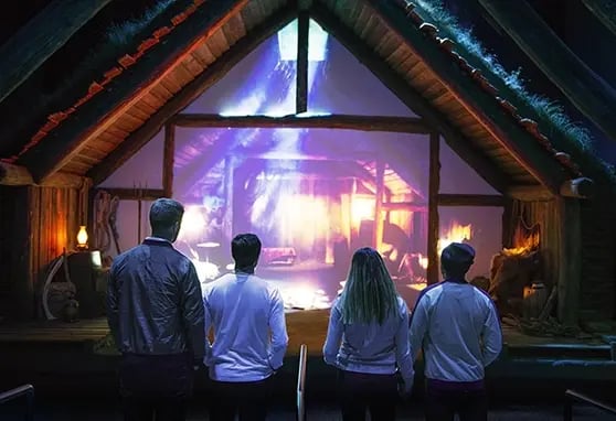 Four visitors stand in front of a vibrant projection of a cozy, historical cabin interior, captivated by the immersive display.