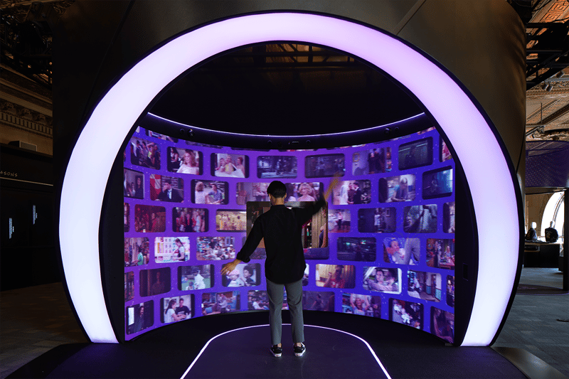 This HBO Max experience immerses visitors in a curved, interactive digital wall surrounded by glowing purple light, allowing them to explore content with dynamic gesture-based controls in a futuristic and visually captivating environment.