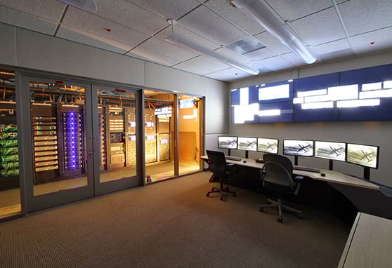 High-tech control room featuring a secure server room with illuminated audiovisual racks, multi-screen operator workstations, and a large video wall display for real-time monitoring and system management at Los Angeles International Airport. 