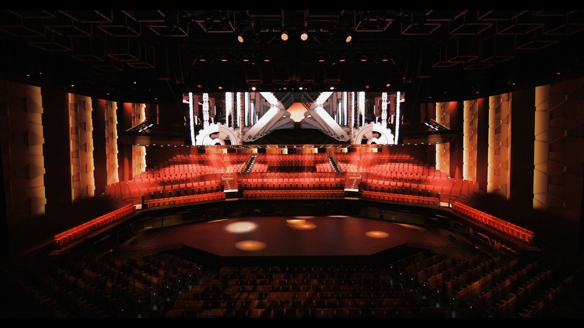 Interior of a futuristic theater with tiered red seating surrounding a central circular stage, geometric wall paneling illuminated with warm lighting, and a large LED wall above the stage displaying a mechanical, industrial-themed graphic.