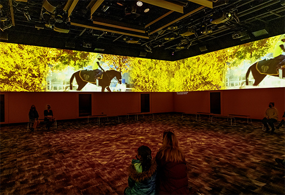 Immersive projection room with wraparound autumn-themed visuals showing a rider on horseback; visitors sit on benches and the floor, surrounded by dynamic ceiling-mounted displays in a darkened, high-tech space