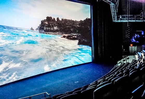 Immersive theater with stadium-style seating and a large curved projection screen displaying an ocean and rocky coastline scene. Blue cushioned chairs are arranged in ascending rows with illuminated aisles with overhead lighting.