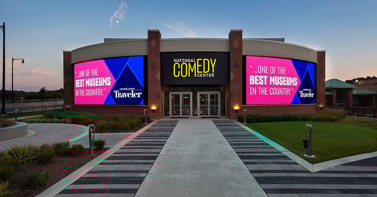 The exterior of the National Comedy Center features dynamic LED displays and digital signage integrated into the building’s facade, showcasing vibrant content related to comedy and upcoming exhibits. These audiovisual elements draw visitors in with animated visuals and engaging messaging, setting a lively tone before entering the museum.