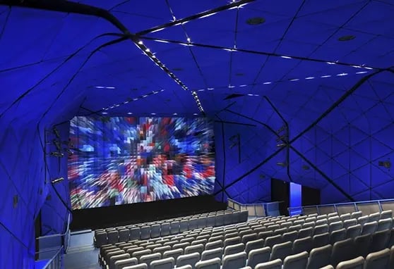 Interior of a modern cinema with a geometric blue design, featuring a bright, vibrant movie screen and empty seating rows.