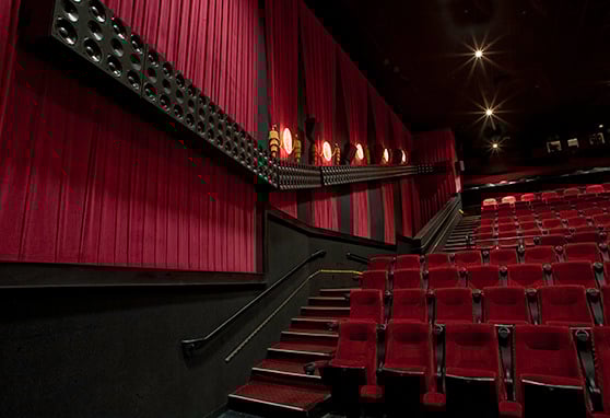 Interior of a movie theater with red velvet seats and matching curtains, angled view showing a tiered seating layout, wall-mounted speakers and soft overhead lighting.