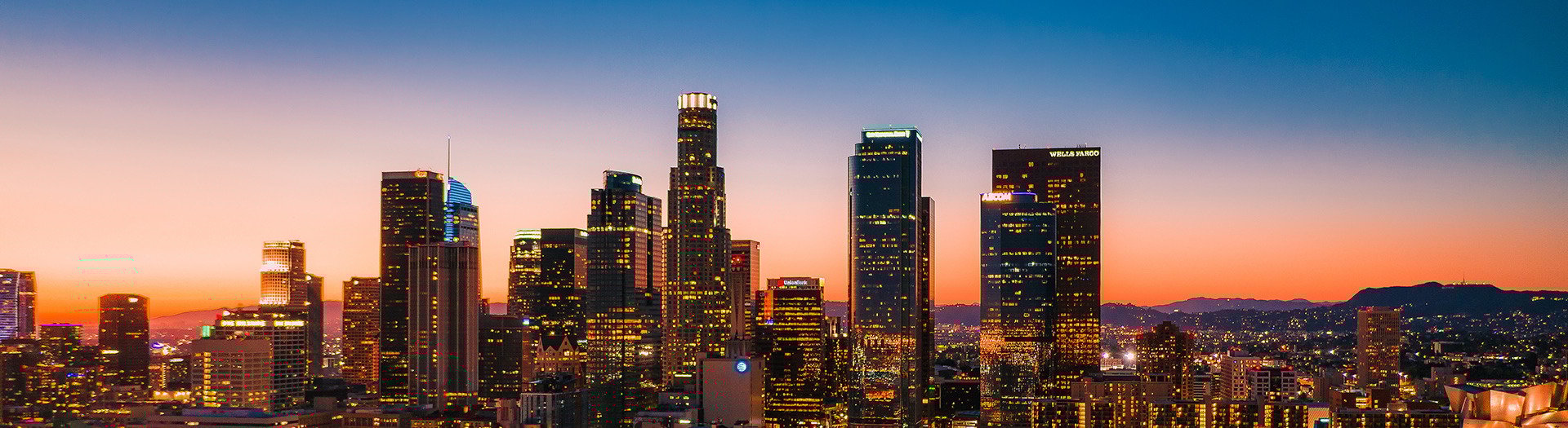 Downtown Los Angeles at dusk is bathed in a warm, golden light as the sun sets behind the skyline, casting long shadows across the city. Skyscrapers begin to glow with interior lights, while traffic and activity add movement to the urban landscape.